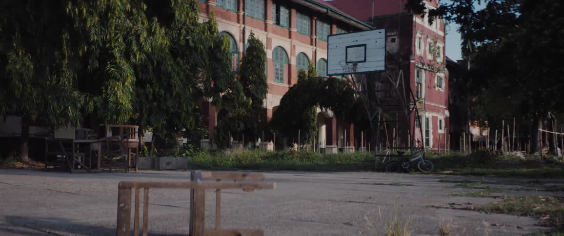 An outdoor basketball court littered with old furniture outside of a dilapidated school building during the day