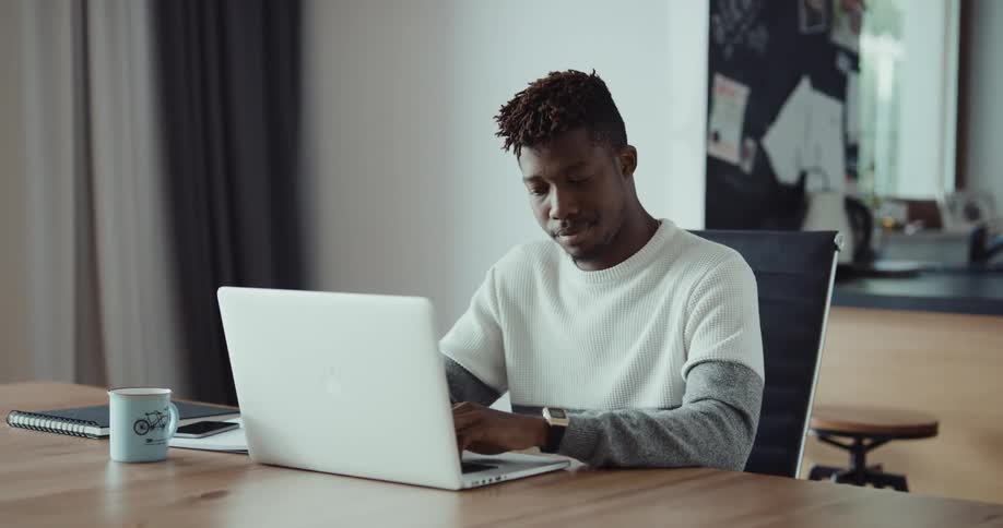 A man is sitting at a table in a home typing on his laptop and looks upset