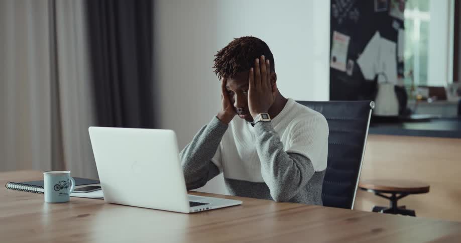A man is sitting at a table typing on a laptop computer and he stops to put his hands on his face and rub his eyes