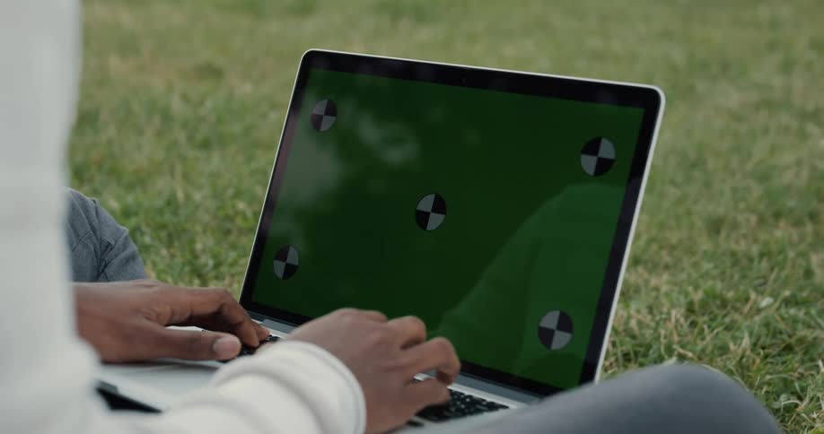 Black man sitting on the grass typing on his laptop on his lap
