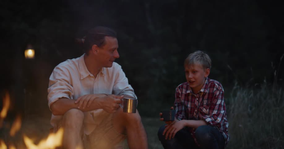 Father with white shirt and brown shorts sits by campfire holding cup next to son talking with
