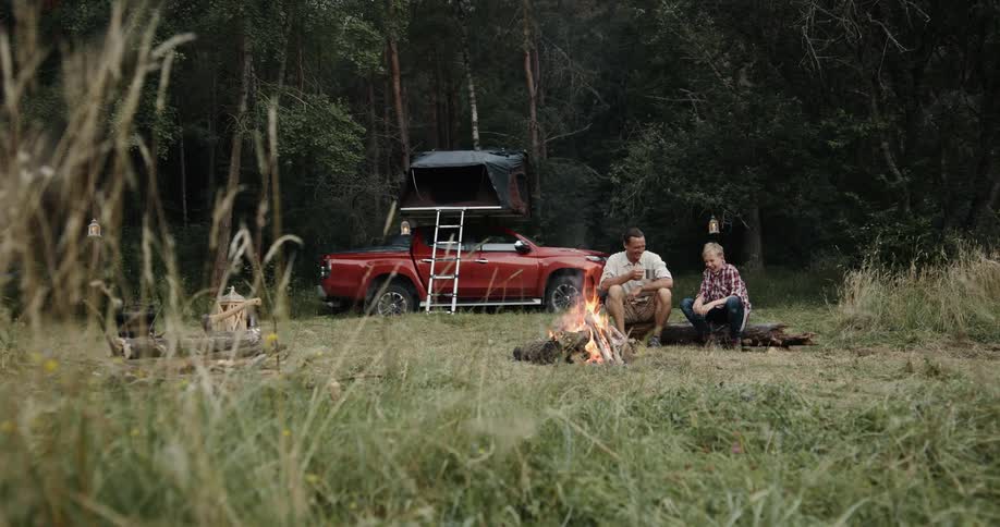 Two males sit at a campfire in a woodland area
