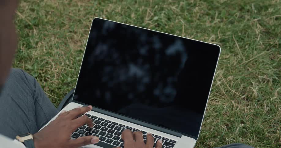 A man types on his computer as he sits on a well manicured lawn