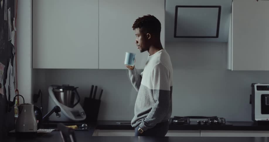 A man stands in his kitchen drinking a cup of coffee then turns and walks away