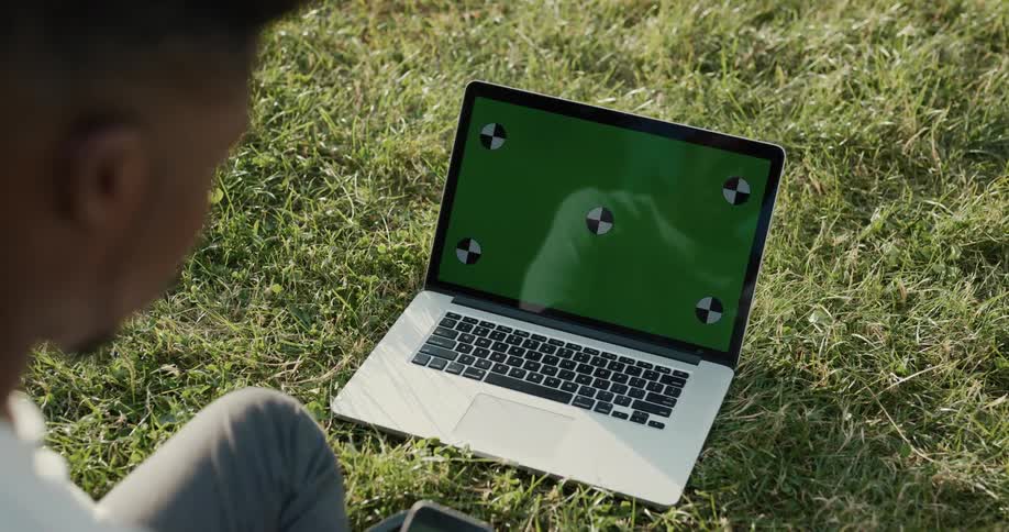 A man sits in the grass before a laptop resting on the ground during the day