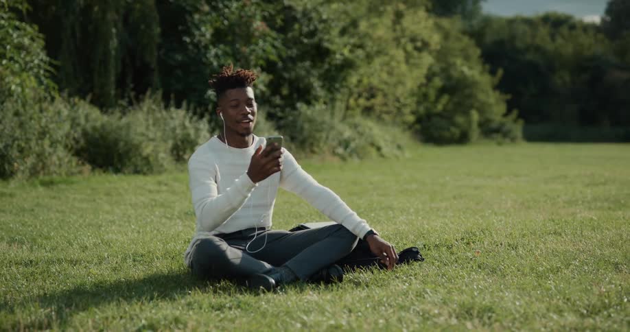 A man is sitting outside in a field near woods talking on his cell phone