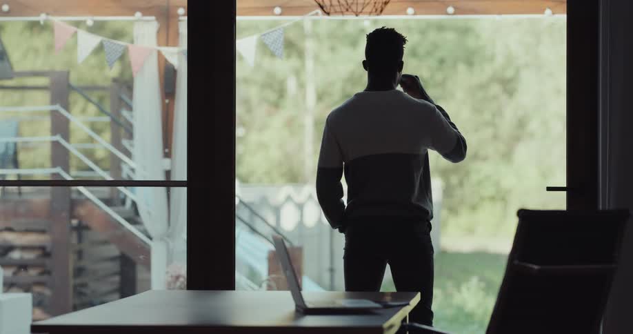 A man sitting before a laptop at a table stands up to gaze out of a nearby window while drinking from a coffee cup