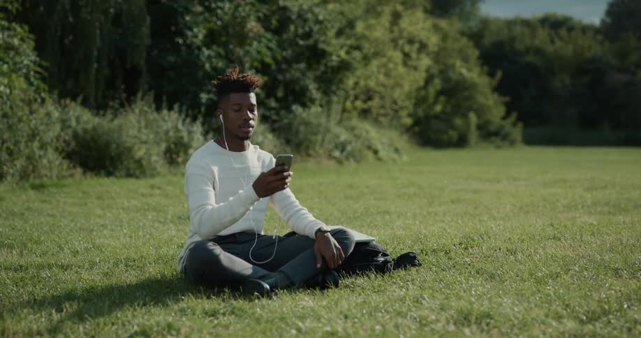 Man with white shirt and green pants sits on lawn wearing earphones and using smartphone