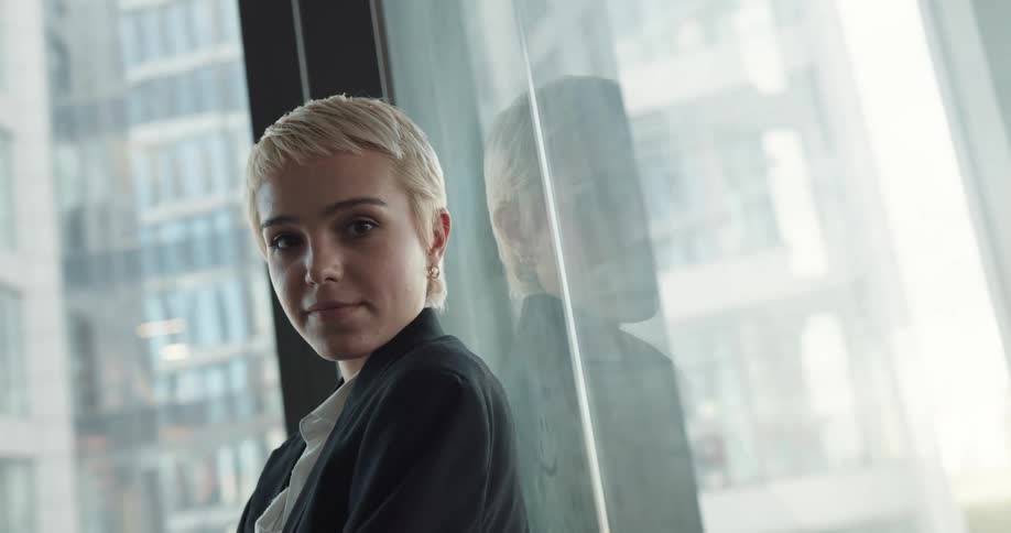 Young blonde female office worker wearing a blazer leaning against a glass window and smiling