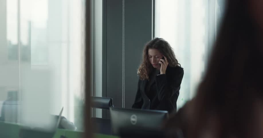 Young female office worker standing near the window in the office talking on the phone