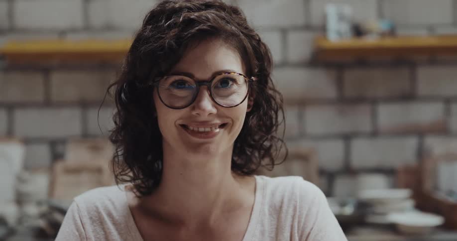 Woman with wavy hair glasses and white shirt looks at camera in the kithcen