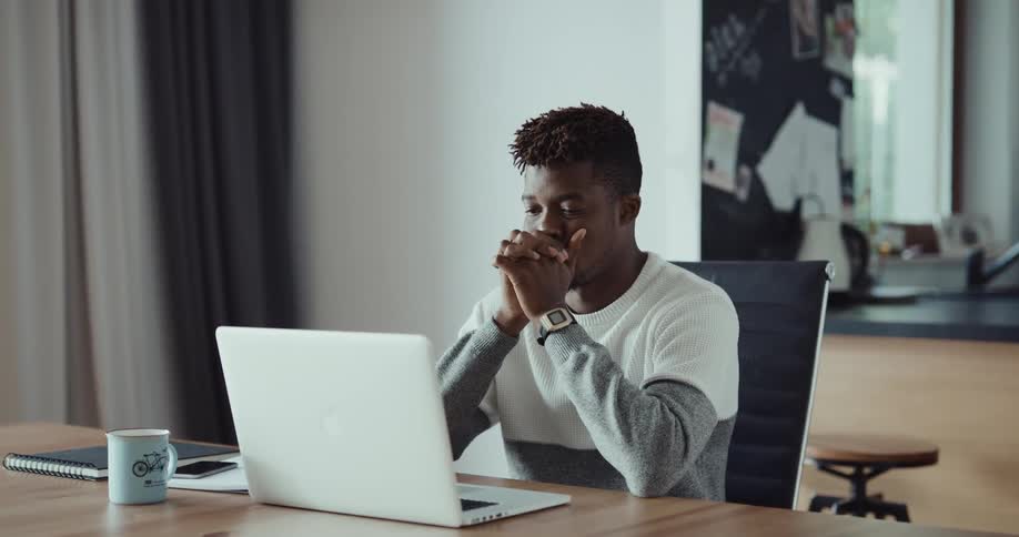 Black man sitting at the desk indoors typing on his laptop and then stopping to think about something