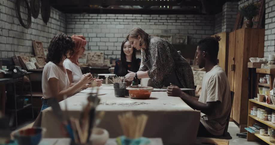 A group of young adults shape together at a table as an older woman cuts clay from a block