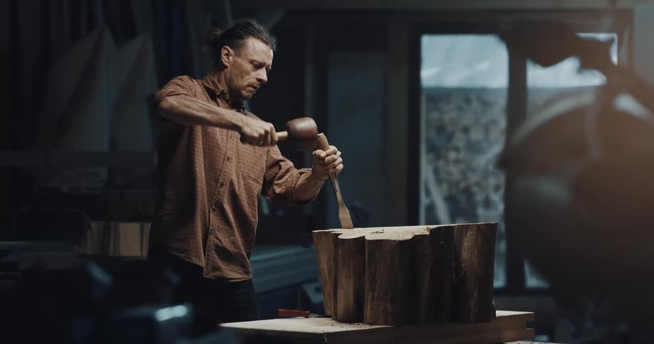 Artisan craftsman carves a tree stump with chisel and hammer inside his workshop