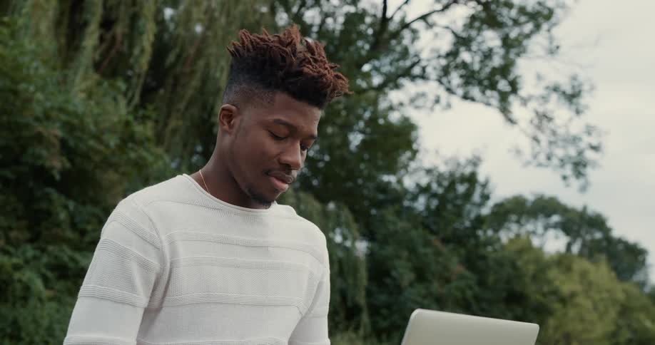 Black man sitting on the grass in the park using his laptop on his lap