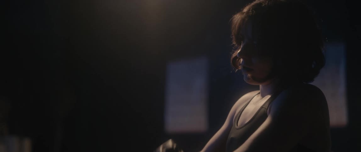 A female fighter sitting on a bench secures her grappling gloves before rising to her feet and walking out of frame