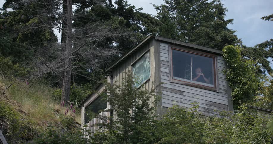 A bearded man with binoculars surveys the landscape from a cabin