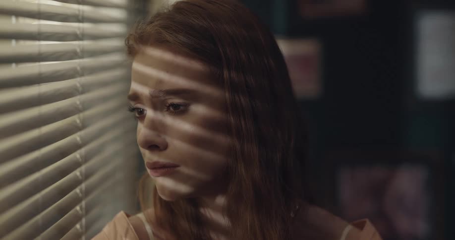 Worried woman looking through the blinds on a window turns to frown at the camera