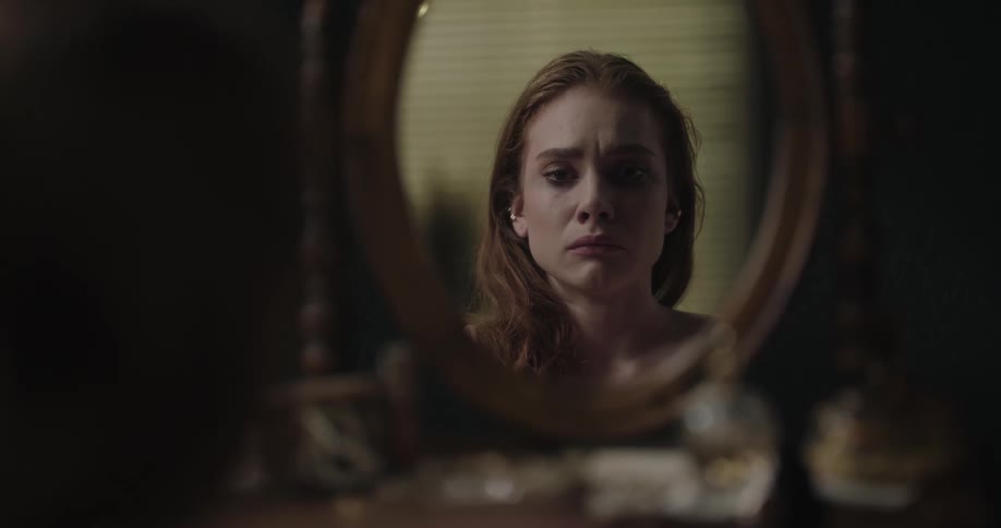An upset woman looks at her reflection in the mirror of a vanity while sitting indoors