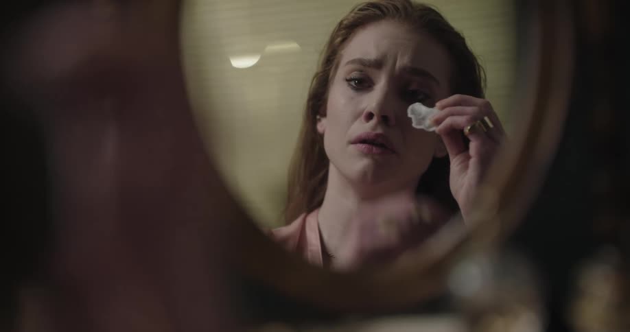 Young redhead woman crying while removing her makeup in front of a mirror