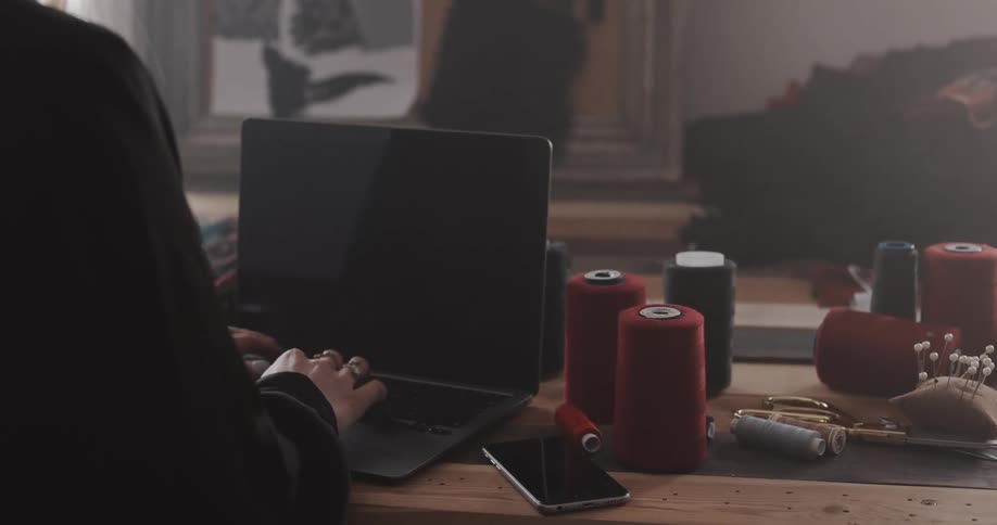 A person types on a laptop while sitting at a table covered in spools of thread