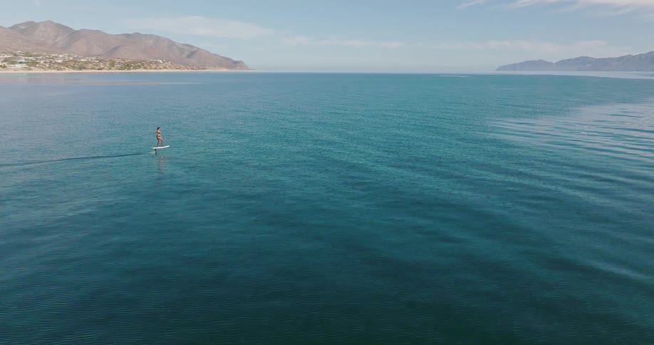 A woman rides an electric hydrofoil surfboard across sparkling clear blue water and there are mountains in the distance