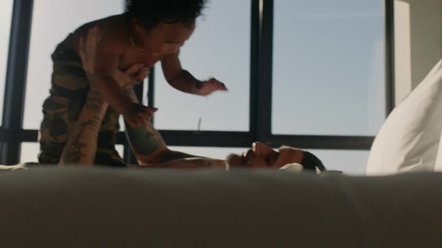 Father lies on his back on bed near window with view of city skyline and plays with his son