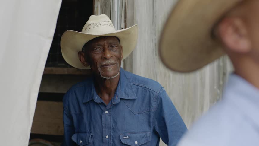 Older man wearing a cowboy hat stands near wall staring