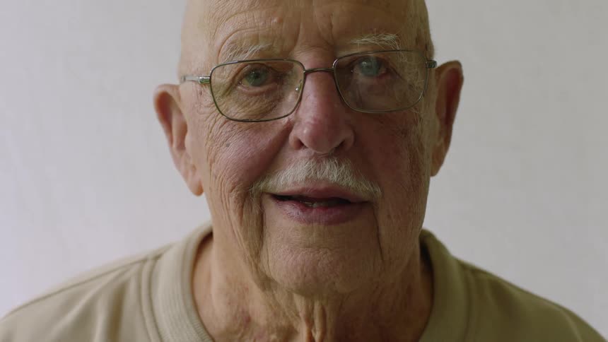 An older man wearing wire frame glasses is looking ahead and smiles