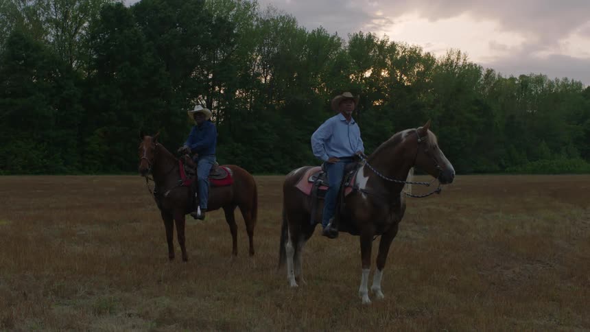 Two cowboys sitting on horses standing in a field near a tree line at sunset