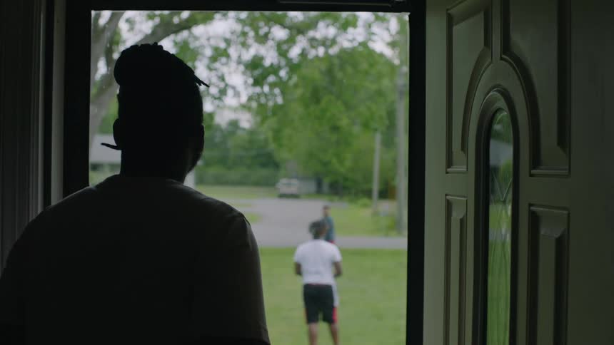A man standing in the doorway watching his sons play catch with a football in the front yard