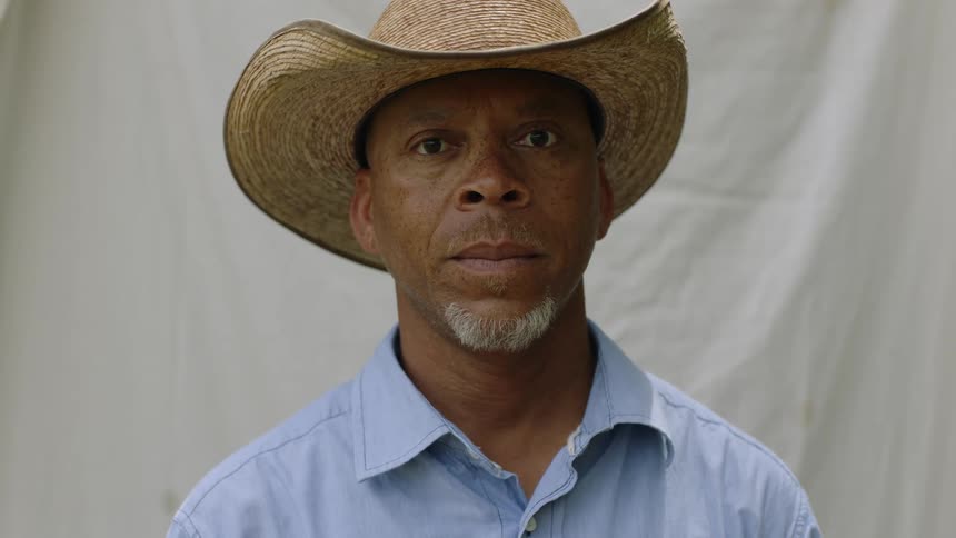 Black man wearing a blue button down shirt and a straw cowboy hat