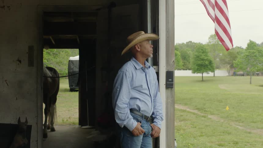 Cowboy reclined on a stable wall under a united states flag looks at the camera