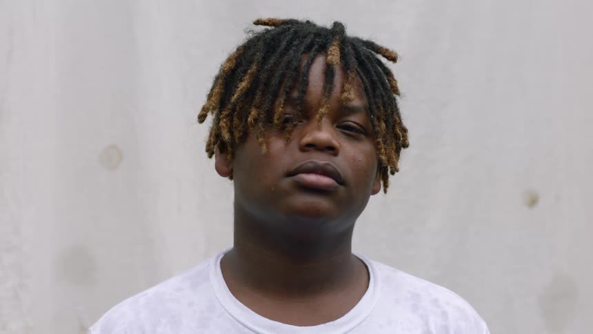 Young boy with dreadlocks stands in front of a white wall looking at the camera