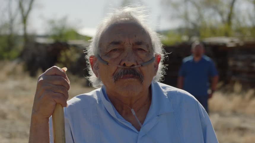 Elderly man leans on a large staff looking at the camera while using a nasal cannula