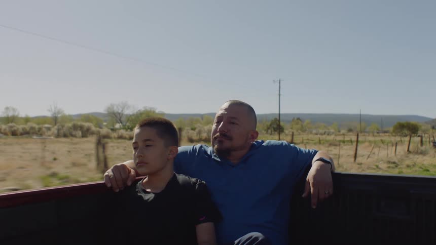 A man and a boy are riding in the back of a pickup truck as it travels on a rural road