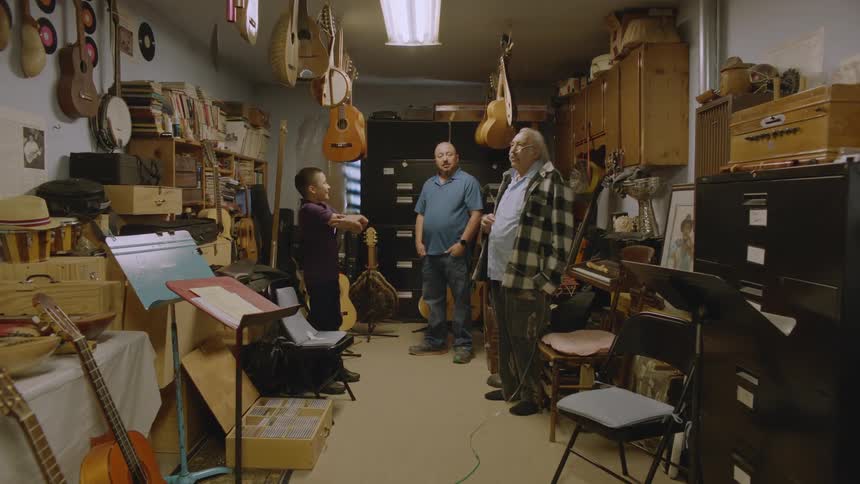 A grandfather standing in a room full of musical instruments talking to his son and grandson