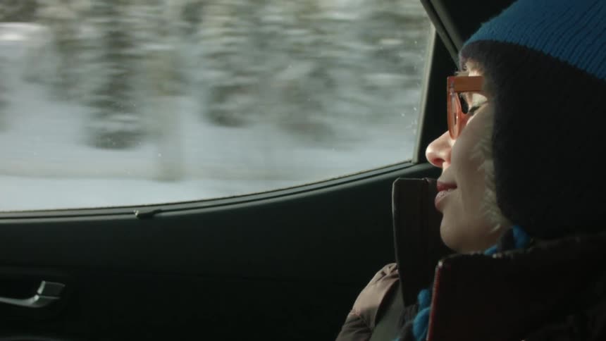 A woman rides in a car through a snowy and wooded area