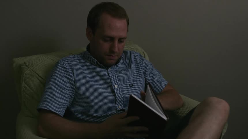 A man sitting in an armchair picks up a book and opens it