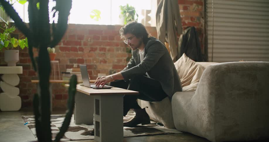 A man types on a laptop while sitting on a sofa indoors during the day