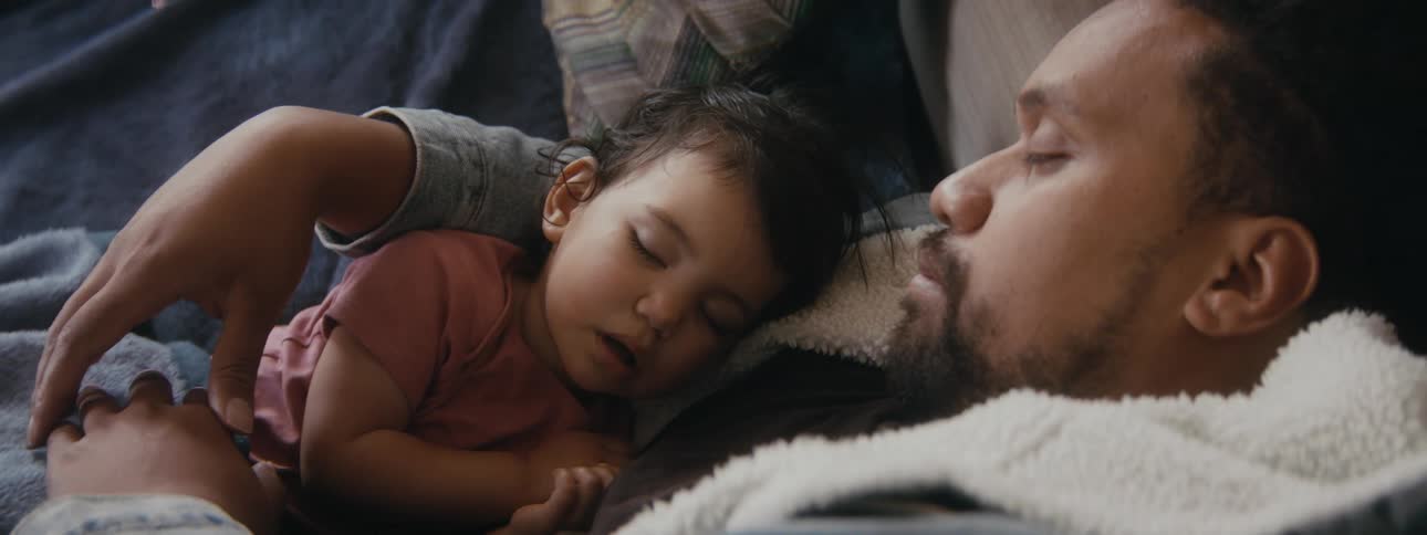 A small child sleeps in the arms of a man sleeping on a sofa