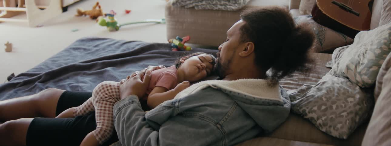 A little girls sleeps on her fathers chest and he raises his head and kisses her head and closes his eyes