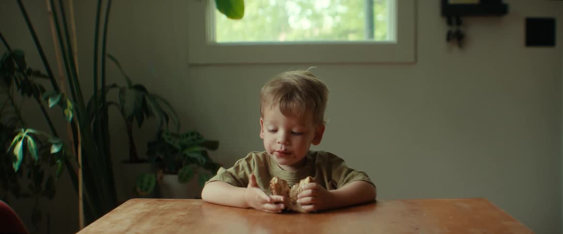 A little boy sitting at the table eating a sandwich licking his hand and fingers