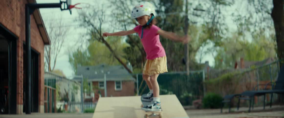 A little girl rides her skateboard up a small ramp in her driveway then turns