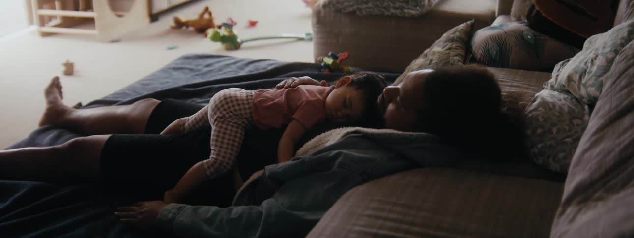 A father sits on the couch and pats his daughters back as she sleeps in his lap