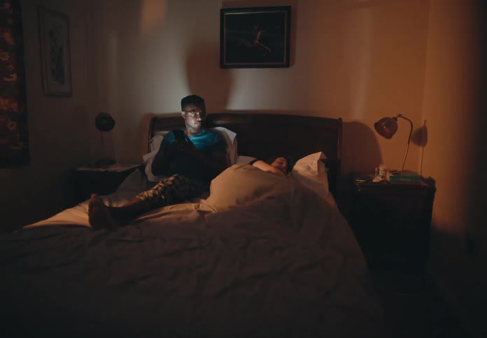A woman is sleeping in bed while her husband sits up watching something on his phone