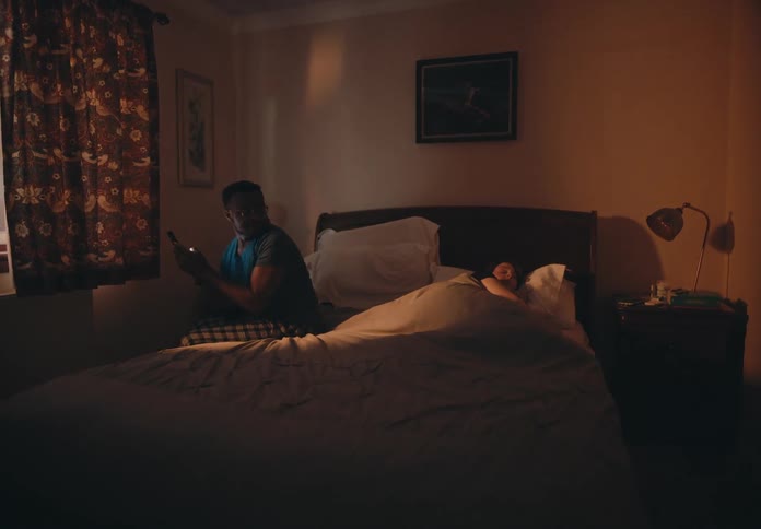 A man sitting on his bed texting on his phone turns and looks over at his sleeping wife