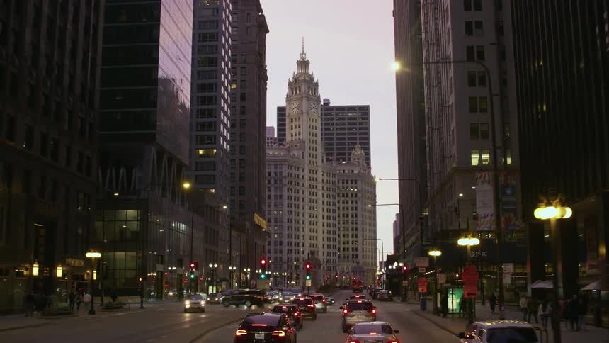 Traffic moves on a busy downtown street lined with tall buildings and people are walking on the sidewalks