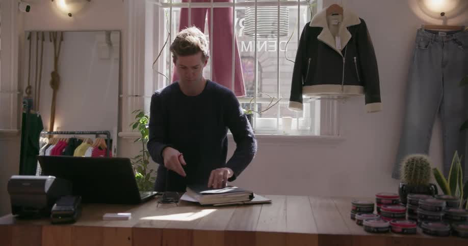 A man behind the counter of a clothing store flips through a binder
