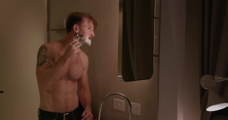 A man is shaving his beard while standing at a bathroom sink